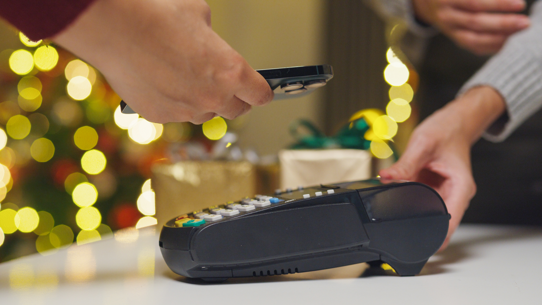 Closeup hand of woman making purchase by cell-phone on electronic payment machine or card reader in small shop with holiday lights in the background.