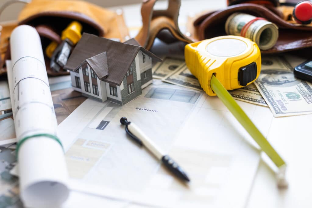 A table with a blueprint, pen, model of a house, measuring tape, and cash on it